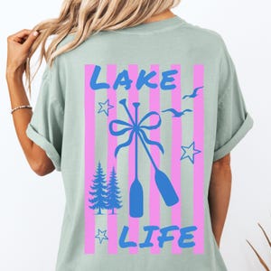 May include: A light green t-shirt with a graphic design on the back. The design features the words "LAKE LIFE" in blue, with pink vertical stripes, oars, stars, birds, and pine trees.