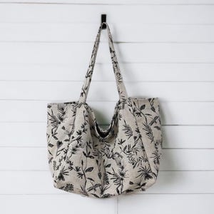 May include: A large, beige tote bag with black floral and dragonfly print. The bag has two long handles and is hanging on a hook.