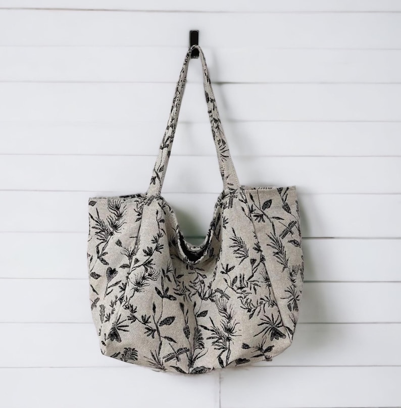 May include: A large, beige tote bag with black floral and dragonfly print. The bag has two long handles and is hanging on a hook.