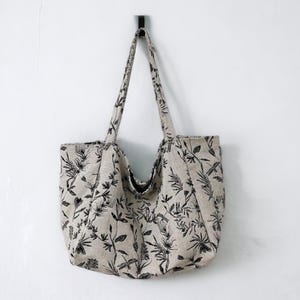 May include: A beige tote bag with black floral and dragonfly print. The bag has two handles and is hanging from a hook.