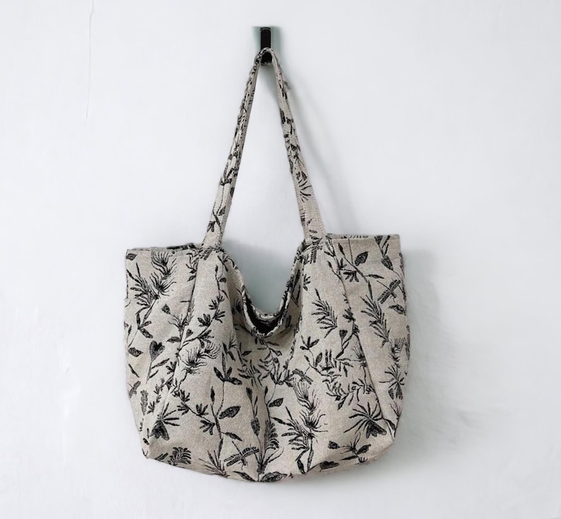May include: A beige tote bag with black floral and dragonfly print. The bag has two handles and is hanging from a hook.