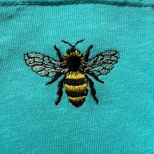 May include: A bright turquoise fabric featuring a detailed embroidered bee at the center. The bee has a black head and thorax, yellow and black striped abdomen, and translucent wings with intricate black stitching details throughout the design.