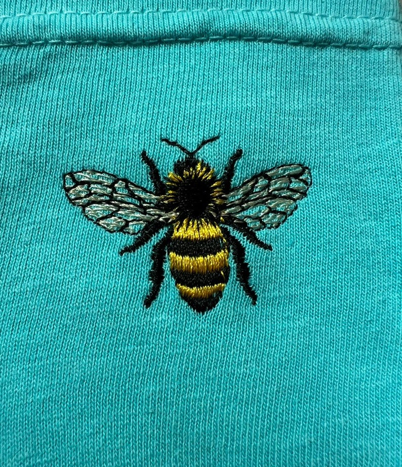 May include: A bright turquoise fabric featuring a detailed embroidered bee at the center. The bee has a black head and thorax, yellow and black striped abdomen, and translucent wings with intricate black stitching details throughout the design.