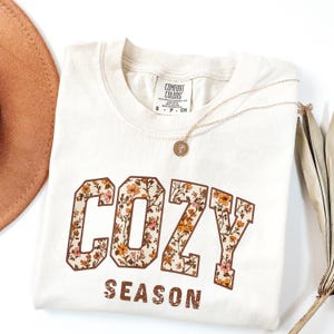 May include: Cream-colored t-shirt with the words "COZY SEASON" in a brown, floral-filled font. A gold necklace with a pendant rests on the shirt. A brown hat and dried plant are visible.