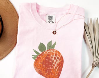 May include: A light pink Comfort Colors t-shirt featuring a large strawberry graphic. The strawberry is red-orange with green leaves. A rose gold necklace with a small pendant rests on the shirt. A brown hat and dried palm leaf are in the background.