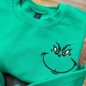 Puede incluir: Una sudadera verde brillante con un rostro del Grinch bordado. La cara del Grinch est&aacute; delineada en negro con ojos amarillos. La sudadera tiene cuello redondo y mangas largas.