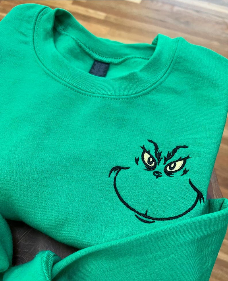 Puede incluir: Una sudadera verde brillante con un rostro del Grinch bordado. La cara del Grinch est&aacute; delineada en negro con ojos amarillos. La sudadera tiene cuello redondo y mangas largas.