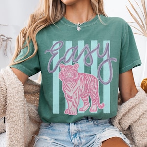 K&ouml;nnte beinhalten: T&uuml;rkisfarbenes T-Shirt mit dem Wort "easy" in Schreibschrift und einer rosa Tigergrafik. Der Tiger ist vor einem Hintergrund aus vertikalen hellblauen und wei&szlig;en Streifen platziert. Das Shirt hat eine lockere Passform.