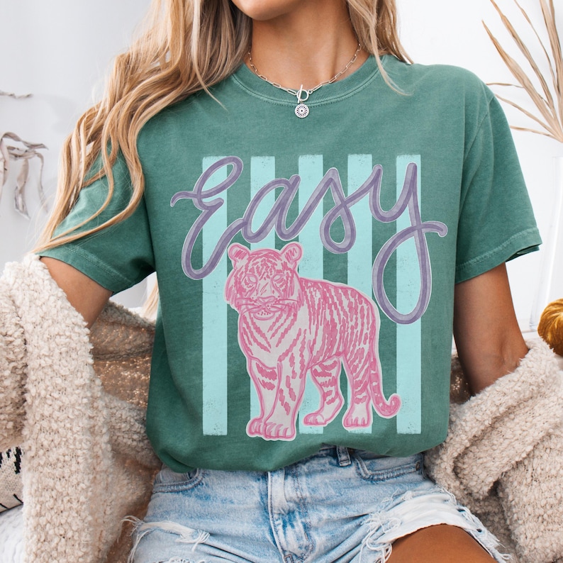 K&ouml;nnte beinhalten: T&uuml;rkisfarbenes T-Shirt mit dem Wort "easy" in Schreibschrift und einer rosa Tigergrafik. Der Tiger ist vor einem Hintergrund aus vertikalen hellblauen und wei&szlig;en Streifen platziert. Das Shirt hat eine lockere Passform.