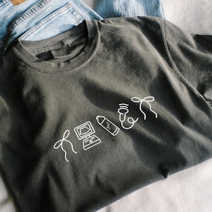 May include: Dark grey t-shirt with a white graphic design featuring a computer, a spray bottle, a microphone, and a bow. The shirt is folded on top of a pair of blue jeans.