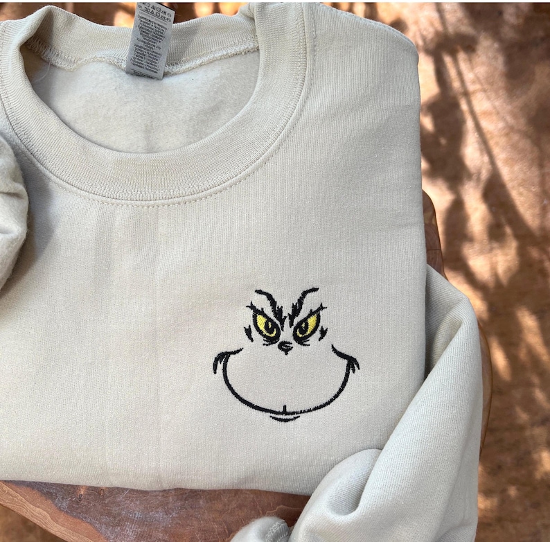 Puede incluir: Una sudadera beige claro con cuello redondo y mangas largas. La parte delantera presenta un dise&ntilde;o bordado de una cara de personaje con ojos amarillos y un contorno negro. La sudadera est&aacute; hecha de un material suave y c&oacute;modo.