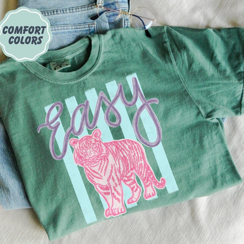K&ouml;nnte beinhalten: Ein gr&uuml;nes T-Shirt mit dem Wort "Easy" in lila Schrift und einer rosa Tigergrafik. Das Shirt hat hellblaue vertikale Streifen hinter dem Wort "Easy" und dem Tiger. Die Worte "Comfort Colors" stehen in einem hellblauen Kreis oben links.