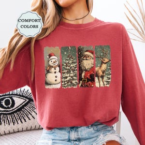 May include: A maroon Comfort Colors sweatshirt featuring a Christmas-themed graphic. The graphic includes a snowman, Christmas tree, Santa Claus, and a reindeer, each in a vertical rectangular panel. The sweatshirt has a crew neck.