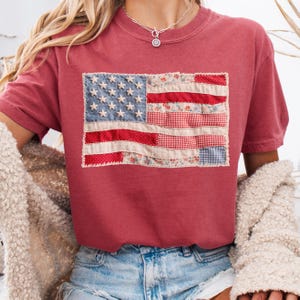 K&ouml;nnte beinhalten: Ein weinrotes T-Shirt mit einem Patchwork-Design der amerikanischen Flagge. Die Flagge hat blaue Sterne, rot-wei&szlig;e Streifen und Blumen- und Vichy-Muster. Das T-Shirt ist aus weichem Material.