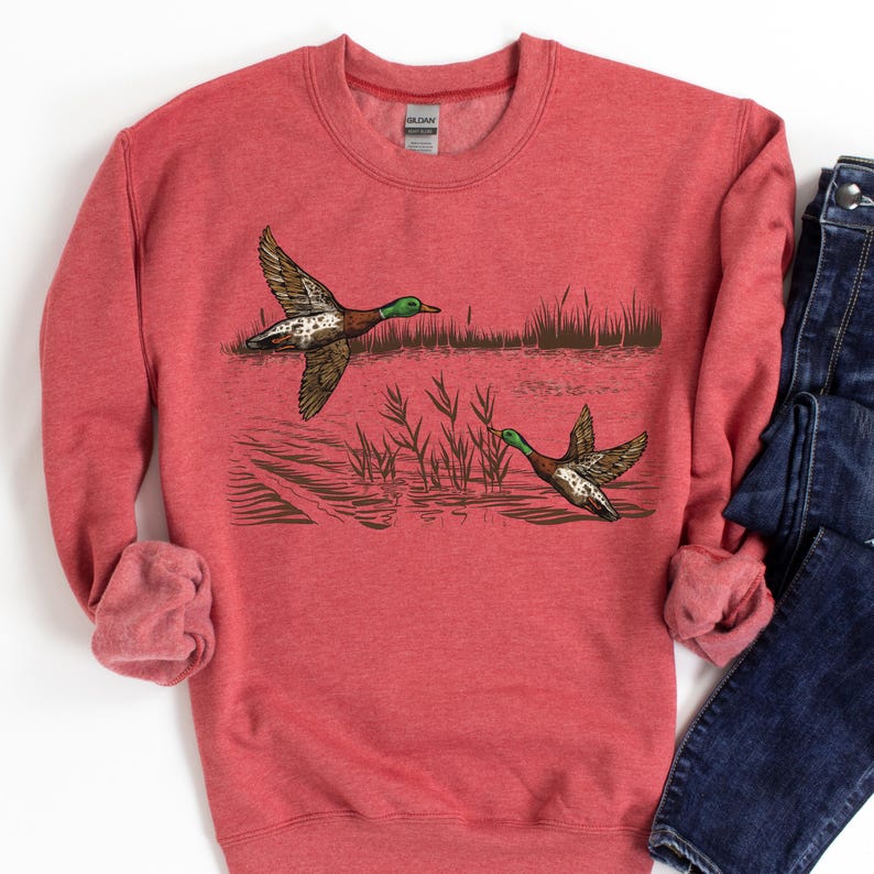 K&ouml;nnte beinhalten: Rotes Sweatshirt mit einer detaillierten Illustration von zwei Enten im Flug &uuml;ber Wasser und Schilf. Die Enten haben gr&uuml;ne K&ouml;pfe, braune Fl&uuml;gel und wei&szlig;e und braune Markierungen. Das Sweatshirt hat einen Rundhalsausschnitt und lange &Auml;rmel.