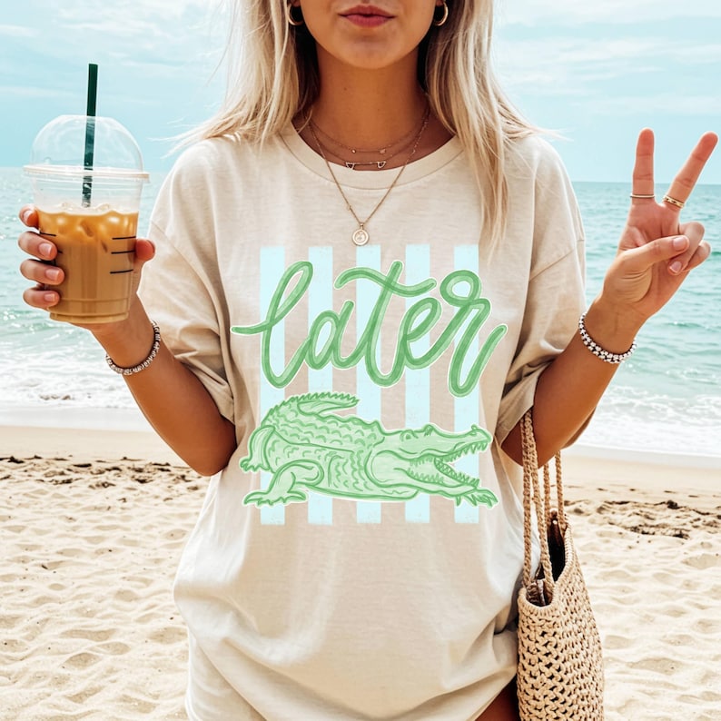 Puede incluir: Camiseta beige con un gr&aacute;fico de cocodrilo verde y la palabra "later" en fuente cursiva. El dise&ntilde;o presenta rayas verticales de color azul claro detr&aacute;s del texto y el cocodrilo. La camiseta se usa en la playa.