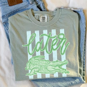 Puede incluir: Camiseta verde salvia con la palabra "later" en fuente cursiva, sobre un dibujo de un caim&aacute;n verde. La camiseta tiene un fondo a rayas y est&aacute; doblada sobre unos vaqueros azul claro.