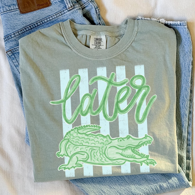 Puede incluir: Camiseta verde salvia con la palabra "later" en fuente cursiva, sobre un dibujo de un caim&aacute;n verde. La camiseta tiene un fondo a rayas y est&aacute; doblada sobre unos vaqueros azul claro.