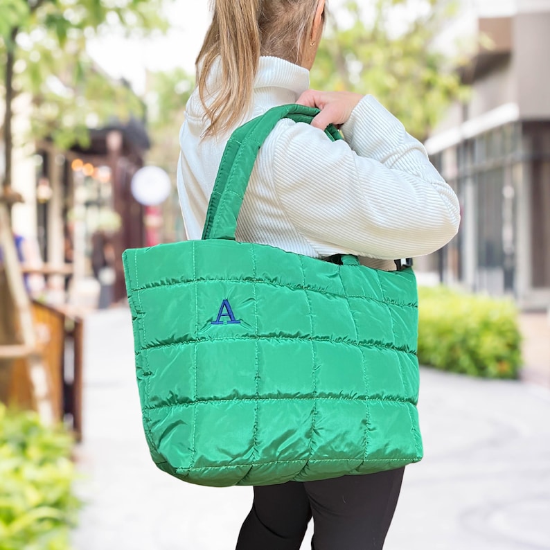 May include: A bright green quilted tote bag with a stitched grid pattern. The bag has a matching green handle and a blue embroidered letter "A" on the front. The bag is carried over the shoulder.