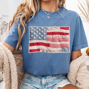 K&ouml;nnte beinhalten: Ein blaues T-Shirt mit einem Patchwork-Design der amerikanischen Flagge. Die Flagge zeigt wei&szlig;e Sterne auf blauem Grund und rot-wei&szlig;e Streifen aus Blumen- und Vichy-Stoffen. Das T-Shirt hat einen l&auml;ssigen Stil.