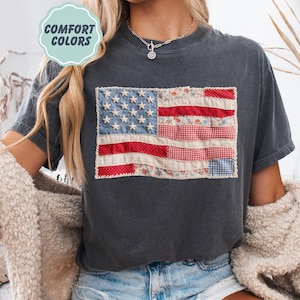 K&ouml;nnte beinhalten: Dunkelgraues T-Shirt mit einem Patchwork-Design der amerikanischen Flagge. Die Flagge verwendet rot-, wei&szlig;- und blau gemusterte Stoffe, darunter Blumen- und Vichymuster. Das T-Shirt hat den Text "COMFORT COLORS" in einem hellgr&uuml;nen Kreis.