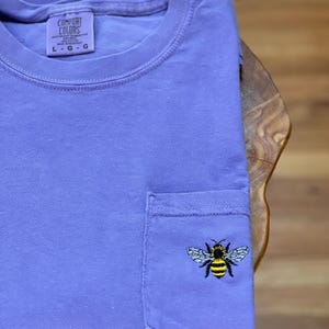 May include: A lavender short-sleeve t-shirt with a crew neckline and a single chest pocket. The pocket features an embroidered bee with yellow and black stripes and white wings. The interior neck label reads Comfort Colors, 100% cotton, size L.