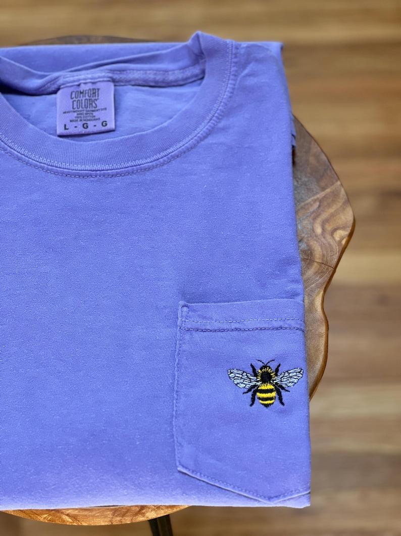 May include: A lavender short-sleeve t-shirt with a crew neckline and a single chest pocket. The pocket features an embroidered bee with yellow and black stripes and white wings. The interior neck label reads Comfort Colors, 100% cotton, size L.