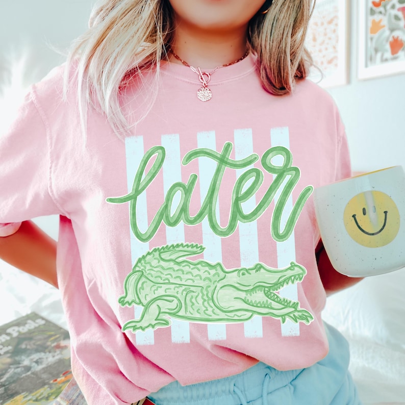 Puede incluir: Camiseta rosa con la palabra "later" en verde, un gr&aacute;fico de cocodrilo verde y rayas blancas verticales. Se sostiene una taza blanca con una carita sonriente amarilla.