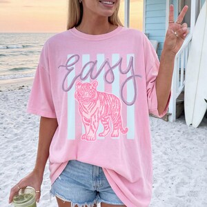 K&ouml;nnte beinhalten: Hellrosa T-Shirt mit dem Wort "Easy" in Schreibschrift &uuml;ber einer rosa Tigerillustration. Der Tiger ist vor einem Hintergrund aus hellblauen vertikalen Streifen platziert. Das Shirt hat kurze &Auml;rmel und eine lockere Passform.