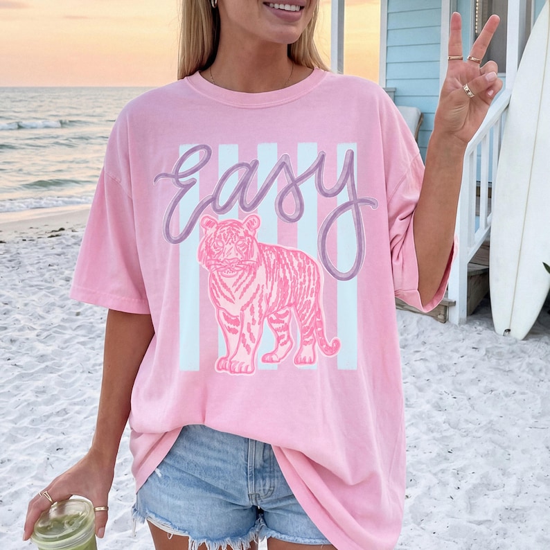 K&ouml;nnte beinhalten: Hellrosa T-Shirt mit dem Wort "Easy" in Schreibschrift &uuml;ber einer rosa Tigerillustration. Der Tiger ist vor einem Hintergrund aus hellblauen vertikalen Streifen platziert. Das Shirt hat kurze &Auml;rmel und eine lockere Passform.
