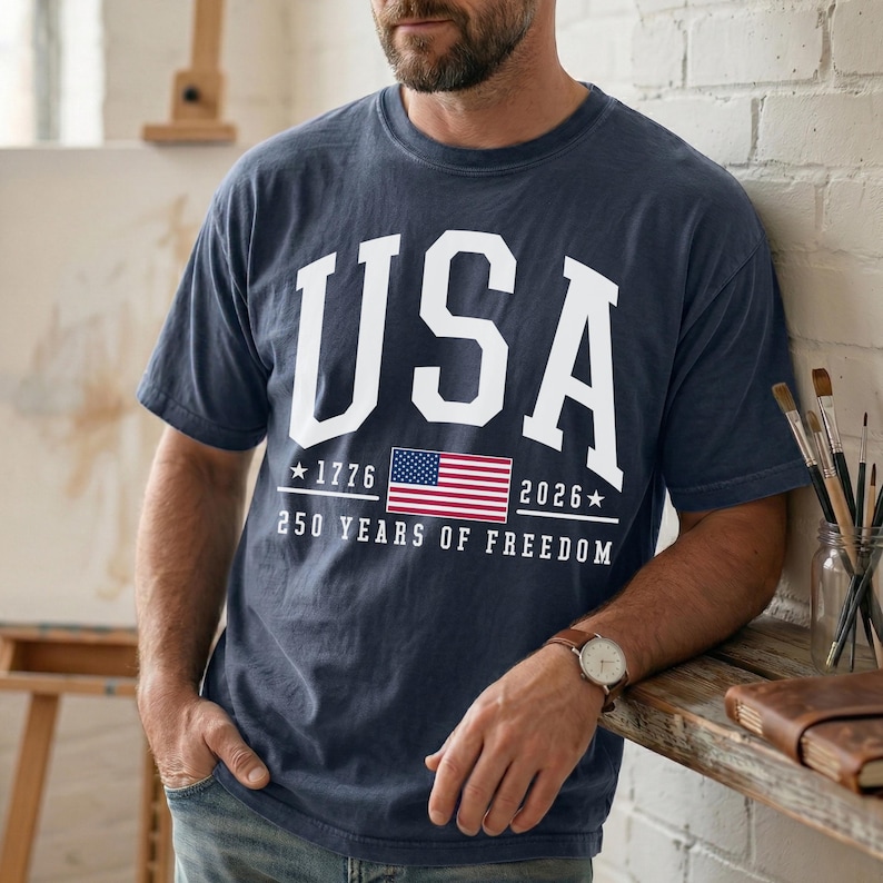 May include: Navy blue t-shirt with the word "USA" in large white block letters. Below the text is the American flag, the years "1776" and "2026", and the phrase "250 YEARS OF FREEDOM".