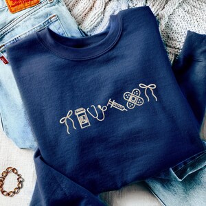 Pode incluir: Camisola azul marinho com s&iacute;mbolos m&eacute;dicos bordados, incluindo um frasco de comprimidos, estetosc&oacute;pio, seringa e penso. A camisola est&aacute; dobrada sobre jeans azuis e uma manta de malha.