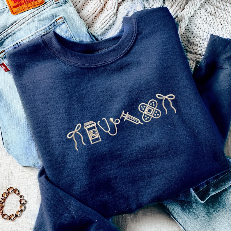 Pode incluir: Camisola azul marinho com s&iacute;mbolos m&eacute;dicos bordados, incluindo um frasco de comprimidos, estetosc&oacute;pio, seringa e penso. A camisola est&aacute; dobrada sobre jeans azuis e uma manta de malha.