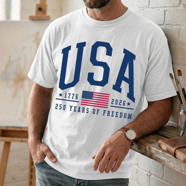 May include: White t-shirt with the word "USA" in navy blue block letters. Below the word "USA" is the American flag, the years 1776 and 2026, and the text "250 YEARS OF FREEDOM". The shirt is short-sleeved.