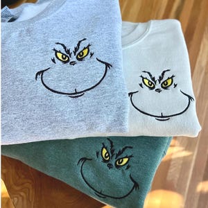 Puede incluir: Tres sudaderas en gris, verde claro y verde azulado, cada una bordada con una cara de dibujos animados con ojos amarillos y una sonrisa negra. Las sudaderas est&aacute;n dobladas y apiladas, mostrando el dise&ntilde;o bordado.
