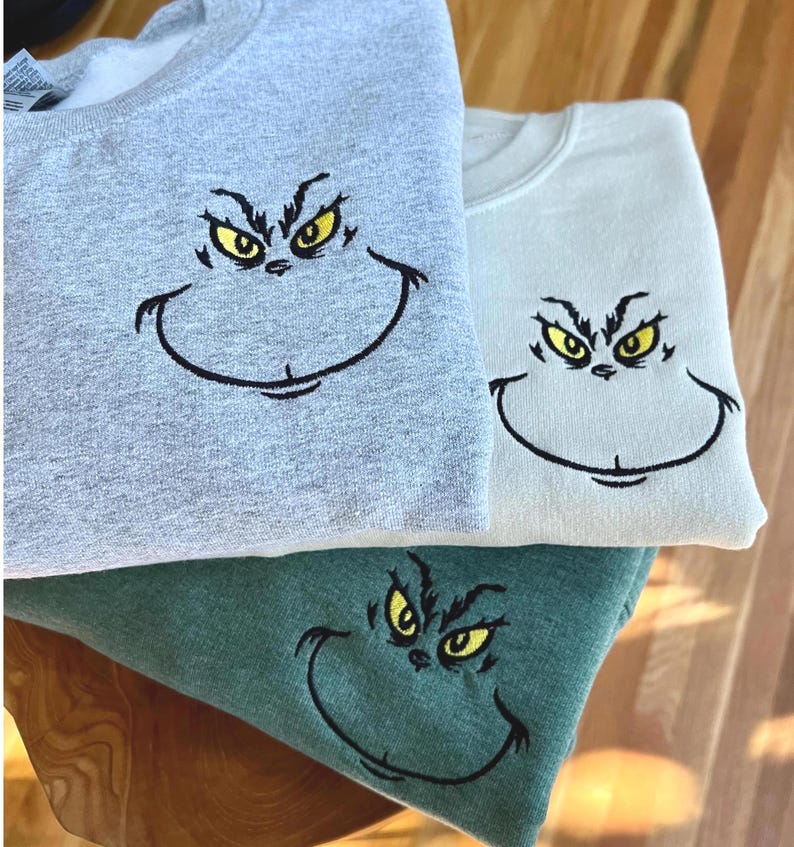 Puede incluir: Tres sudaderas en gris, verde claro y verde azulado, cada una bordada con una cara de dibujos animados con ojos amarillos y una sonrisa negra. Las sudaderas est&aacute;n dobladas y apiladas, mostrando el dise&ntilde;o bordado.
