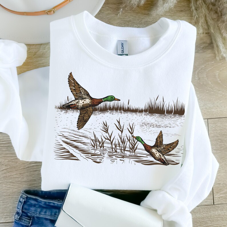 K&ouml;nnte beinhalten: Wei&szlig;es Sweatshirt mit einer detaillierten Illustration von zwei Enten im Flug &uuml;ber Wasser und Schilf. Die Enten sind braun, gr&uuml;n und wei&szlig;. Das Sweatshirt ist aus einem weichen Material.