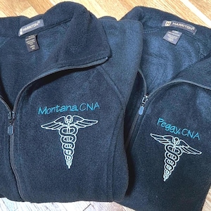 May include: Two black fleece zip-up jackets with embroidered medical caduceus symbols. One jacket says "Montana, CNA" in teal, the other says "Peggy, CNA". Both jackets have the "HARRITON" brand label.