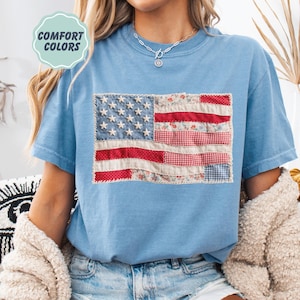 K&ouml;nnte beinhalten: Hellblaues T-Shirt mit einem Patchwork-Design der amerikanischen Flagge. Die Flagge verwendet Stoffe mit roten, wei&szlig;en und blauen Mustern. Das Shirt hat den Text "COMFORT COLORS" in einem hellgr&uuml;nen Kreis.