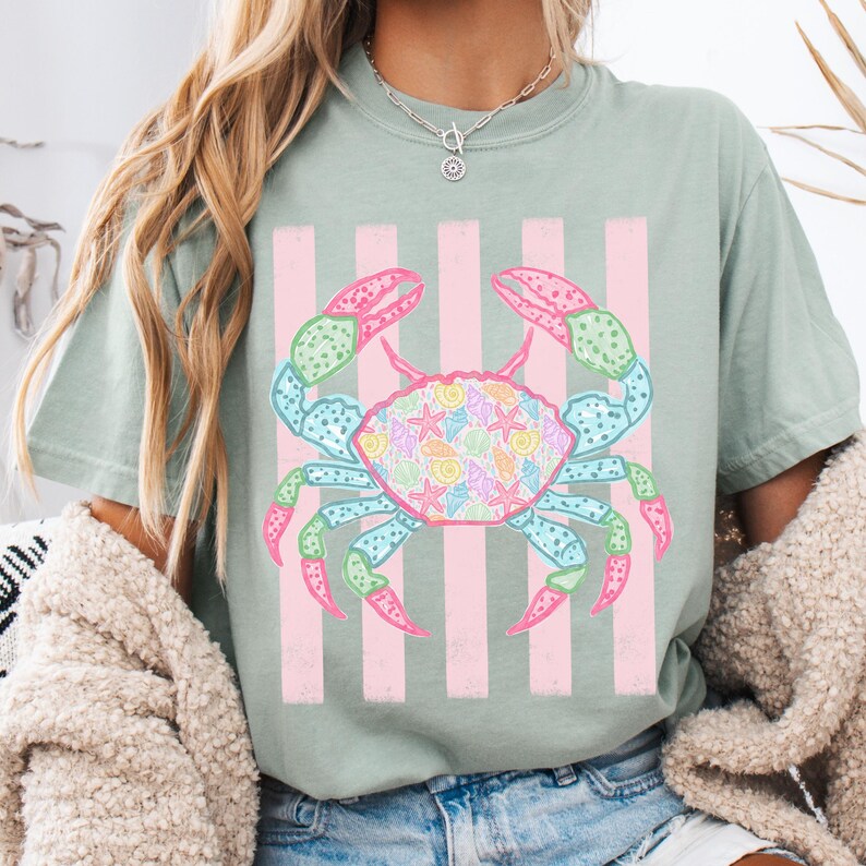 May include: A light green t-shirt featuring a colorful crab design. The crab has pink, blue, and green accents, with a shell pattern of seashells and starfish. Pink vertical stripes are behind the crab.