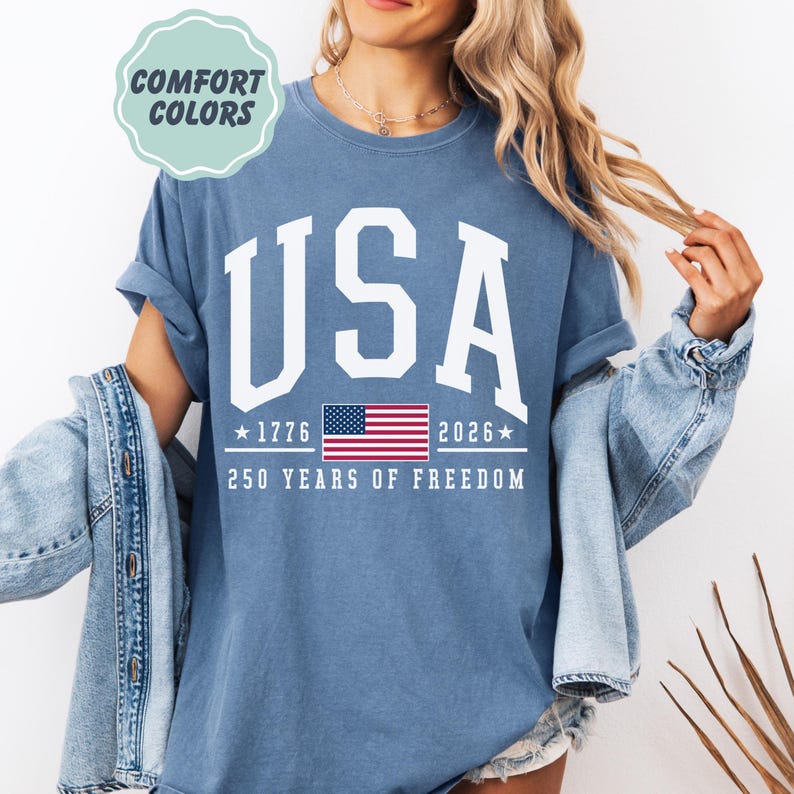 May include: A blue t-shirt with the word "USA" in large white letters. Below the word "USA" is the American flag, the years 1776 and 2026, and the text "250 YEARS OF FREEDOM". The shirt has rolled-up sleeves and a relaxed fit.