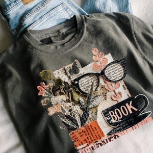 May include: A dark gray t-shirt with a collage design featuring glasses, flowers, and the word "BOOK." Light blue denim jeans are visible in the background. The design incorporates vintage paper elements and a coffee cup.
