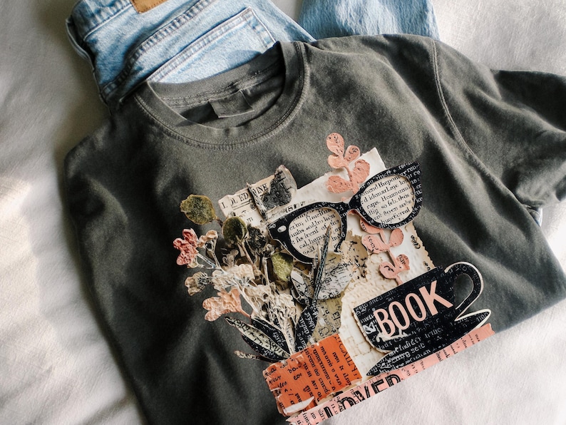 May include: A dark gray t-shirt with a collage design featuring glasses, flowers, and the word "BOOK." Light blue denim jeans are visible in the background. The design incorporates vintage paper elements and a coffee cup.