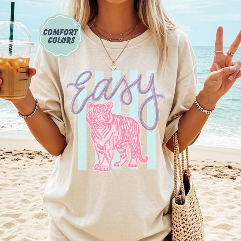 K&ouml;nnte beinhalten: Beiges T-Shirt mit dem Wort "Easy" in lavendelfarbener Schrift &uuml;ber einer rosa Tiger-Illustration. Der Tiger ist vor einem Hintergrund aus hellblauen vertikalen Streifen platziert. Das Shirt hat auch ein "Comfort Colors"-Label.