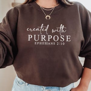 May include: A dark brown crew neck sweatshirt featuring the white text "created with PURPOSE" and "EPHESIANS 2:10". The sweatshirt is a casual style.