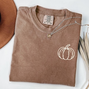 May include: A brown short-sleeve t-shirt with a crew neckline and a white line-art pumpkin graphic on the chest. The shirt features a Comfort Colors brand label inside the collar and is paired with a delicate silver-tone chain necklace.