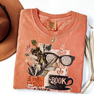 May include: Coral-colored Comfort Colors t-shirt with a collage design featuring glasses, a coffee cup with the word "BOOK", and floral elements. A gold necklace and a brown hat are also visible.