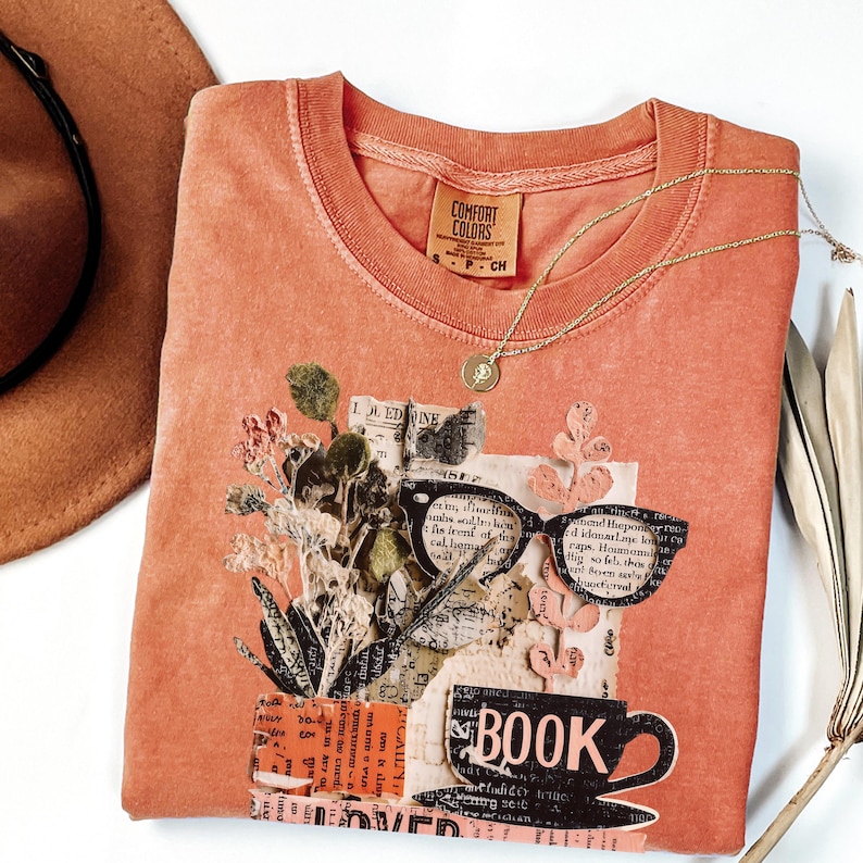 May include: Coral-colored Comfort Colors t-shirt with a collage design featuring glasses, a coffee cup with the word "BOOK", and floral elements. A gold necklace and a brown hat are also visible.