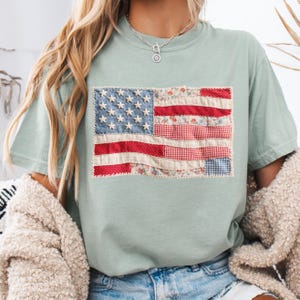 K&ouml;nnte beinhalten: Ein hellgr&uuml;nes T-Shirt mit einem Patchwork-Design der amerikanischen Flagge. Die Flagge hat ein blaues Denim-Feld mit wei&szlig;en Sternen, rot-wei&szlig; gestreifte Abschnitte sowie Blumen- und Vichy-Muster. Das T-Shirt ist aus weichem Material.