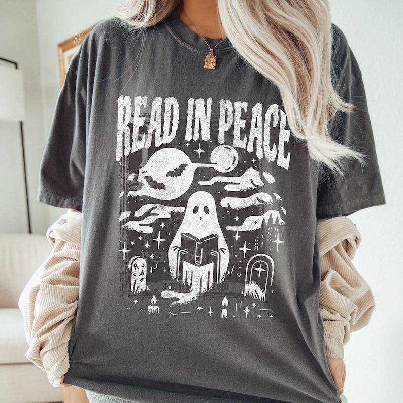 May include: Dark gray t-shirt with a white graphic that reads "READ IN PEACE." The graphic features a ghost reading a book in a graveyard setting with a full moon, bats, and candles. A spooky, Halloween-themed design.